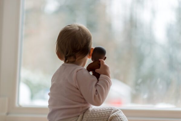 Little baby girl hugging a toy watching the winter snow outdoors as she sits in front of a large view window.